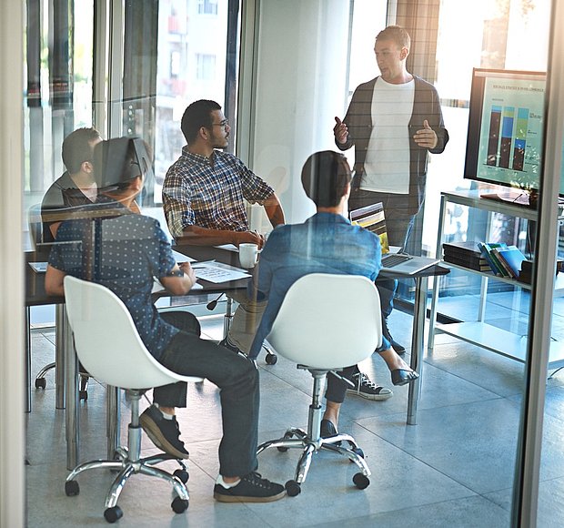 ine Gruppe von Mitarbeitern, die sich in einem Großraumbüro versammelt hat, um ein Projekt zu besprechen. Der Raum verfügt über große Fenster mit Stadtblick und einen Computerbildschirm, auf dem Datendiagramme angezeigt werden. Bei dem Treffen blicken zwei Personen zu einem Mann auf, der steht und zur Gruppe spricht, während die anderen aufmerksam zuhören.