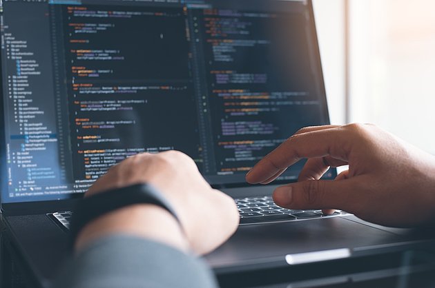 Nahaufnahme einer Person, die auf einer Laptoptastatur tippt. Die Person sitzt an einem Schreibtisch vor einem Laptop, auf dessen Bildschirm verschiedene Zeilen Programmcode zu sehen sind. Die Hände der Person befinden sich auf der Tastatur, wobei eine Hand den Laptop am Gehäuserand hält. Das Bild fängt Details der Hände, der Tastatur und des Bildschirms mit dem Code ein.