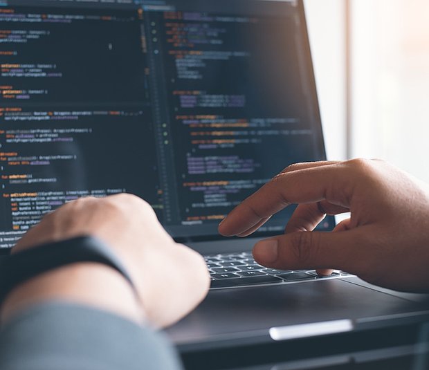 Nahaufnahme einer Person, die auf einer Laptoptastatur tippt. Die Person sitzt an einem Schreibtisch vor einem Laptop, auf dessen Bildschirm verschiedene Zeilen Programmcode zu sehen sind. Die Hände der Person befinden sich auf der Tastatur, wobei eine Hand den Laptop am Gehäuserand hält. Das Bild fängt Details der Hände, der Tastatur und des Bildschirms mit dem Code ein.